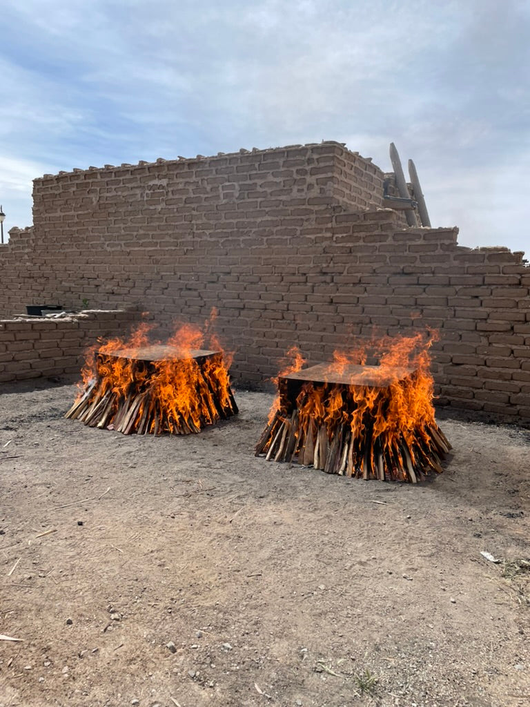 In broad daylight, two square fire pits burn with surrounding flames in front of an adobe brick wall at Poeh Cultural Center